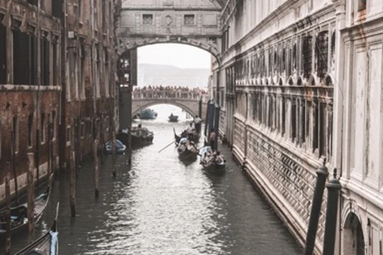 Small boats occupy a narrow canal between tall weathered buildings, with two bridges spanning the water ahead.