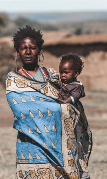 An adult wearing a blue patterned wrap holds a child, with blurred buildings and bare ground behind them.